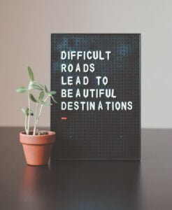 A plant in a pot sitting next a small board with text: "Difficult roads lead to beautiful destinations"
