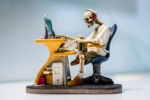 A quirky skeleton figurine wearing headphones sits at a desk with a computer, symbolizing burnout.